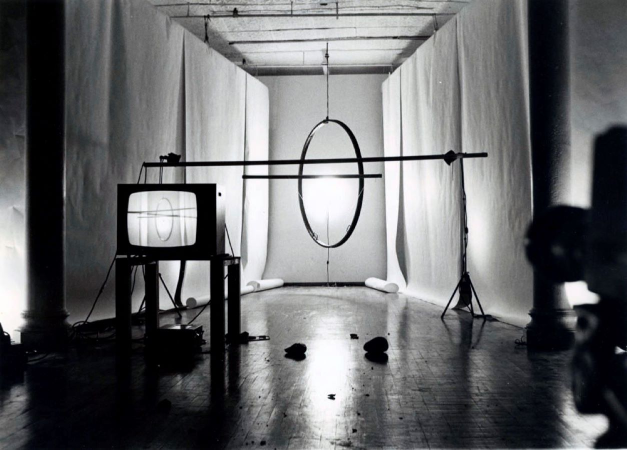 A metal hoop and two metal beams hang from the ceiling, surrounded by white backdrops. In the foreground is a TV monitor on a stand displaying an image of the hoop and beams.