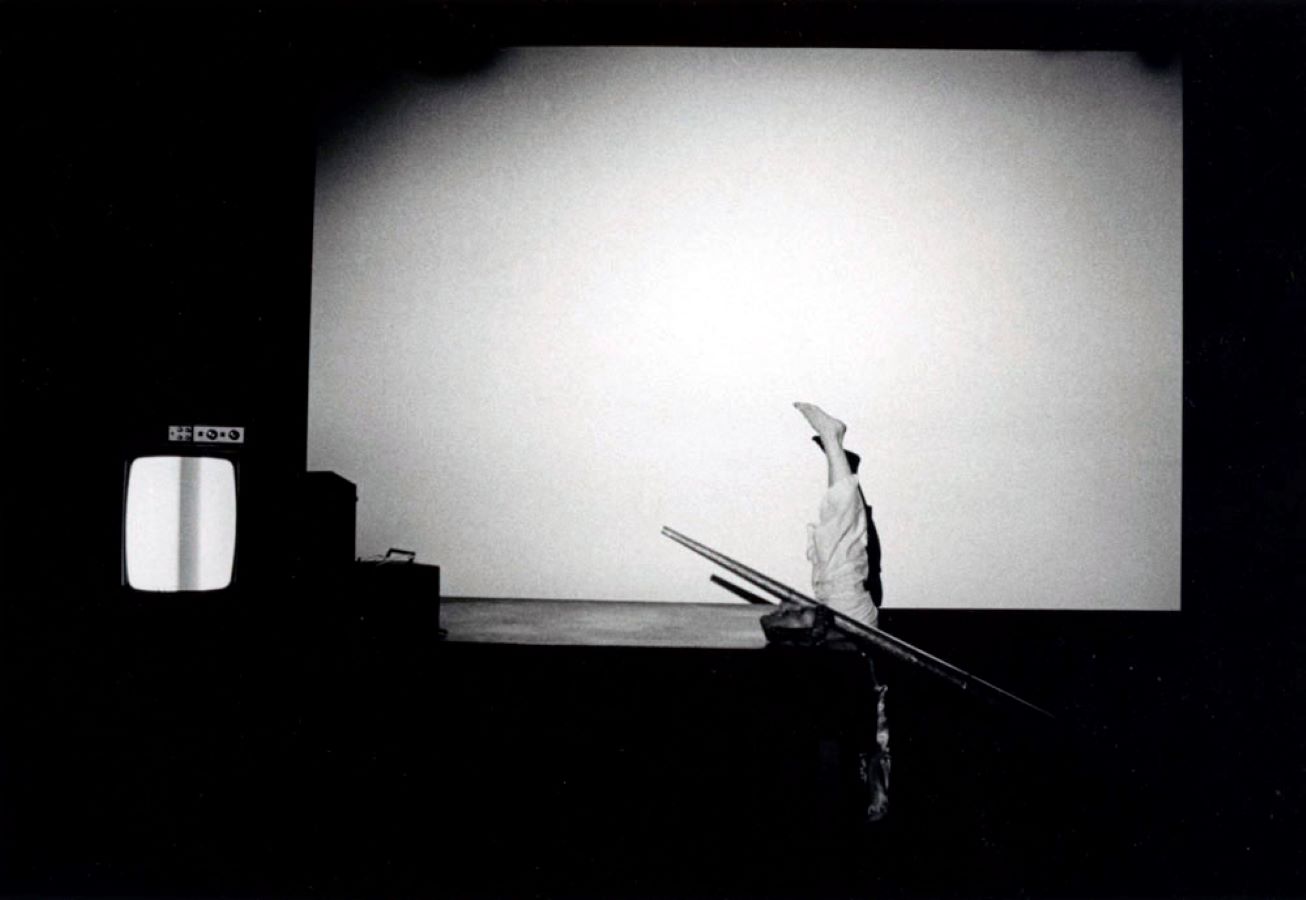 Jonas doing a handstand in between a blank screen and a table with a large metal cone, a mask, and a bag on top; to the left is a TV monitor.