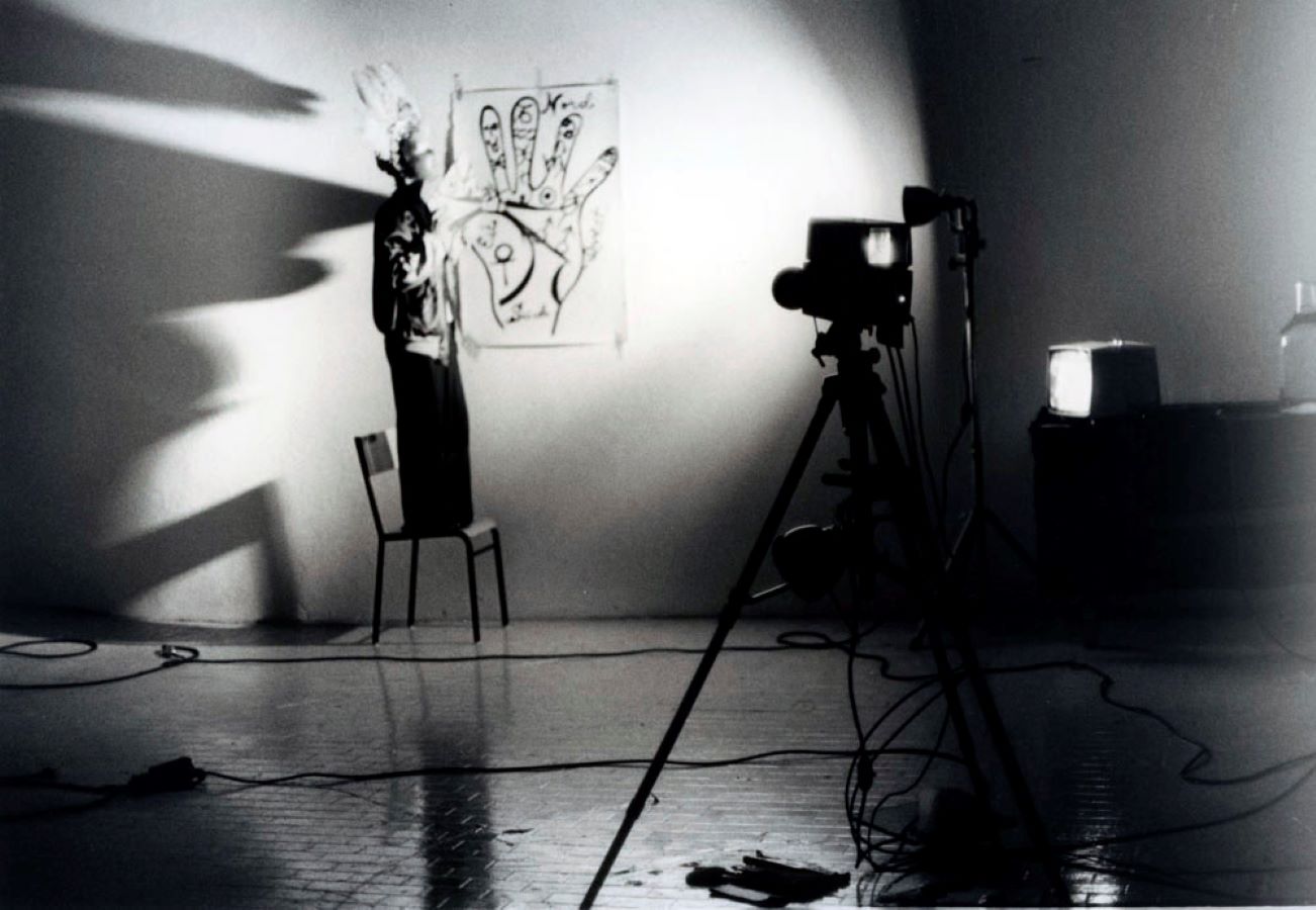 Jonas, wearing a mask and feathered headpiece, stands on a chair next to a drawing of a hand taped to the wall. There is a light pointing at Jonas, which casts dramatic shadows, and a camera on a tripod.