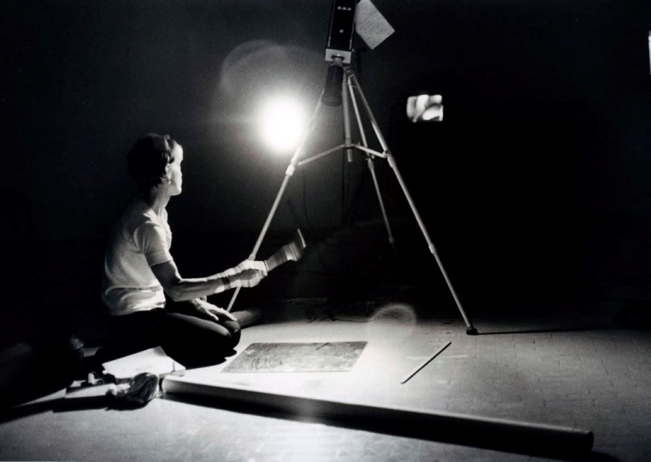 Jonas kneels below a camera on a tripod with a spoon in her right hand