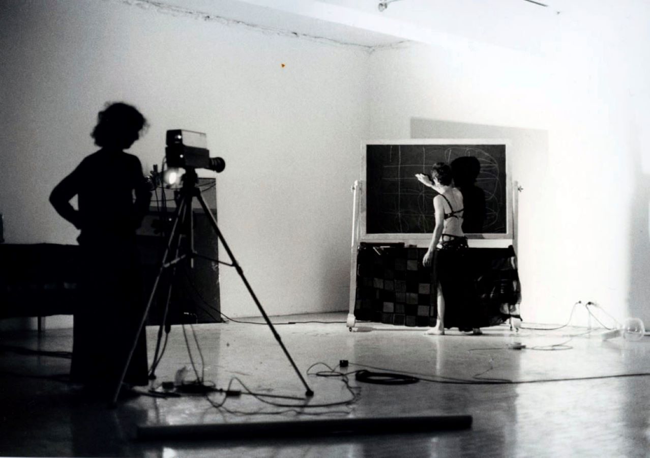 A camerawoman stands behind a tripod while Jonas draws on a blackboard