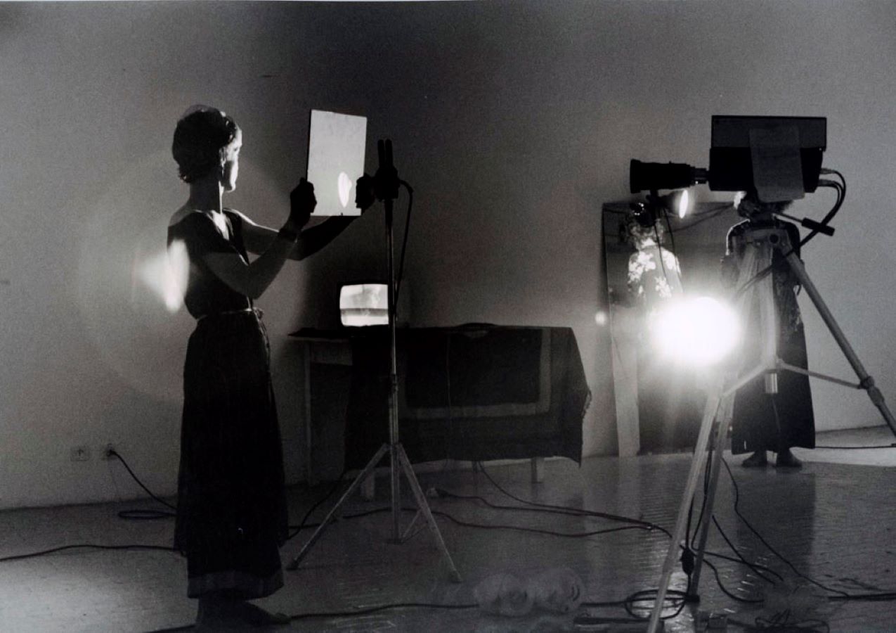 Jonas stands in front of a camera on a tripod while holding up a mirror. In the background is a TV monitor on a table, a large mirror, and a camerawoman.