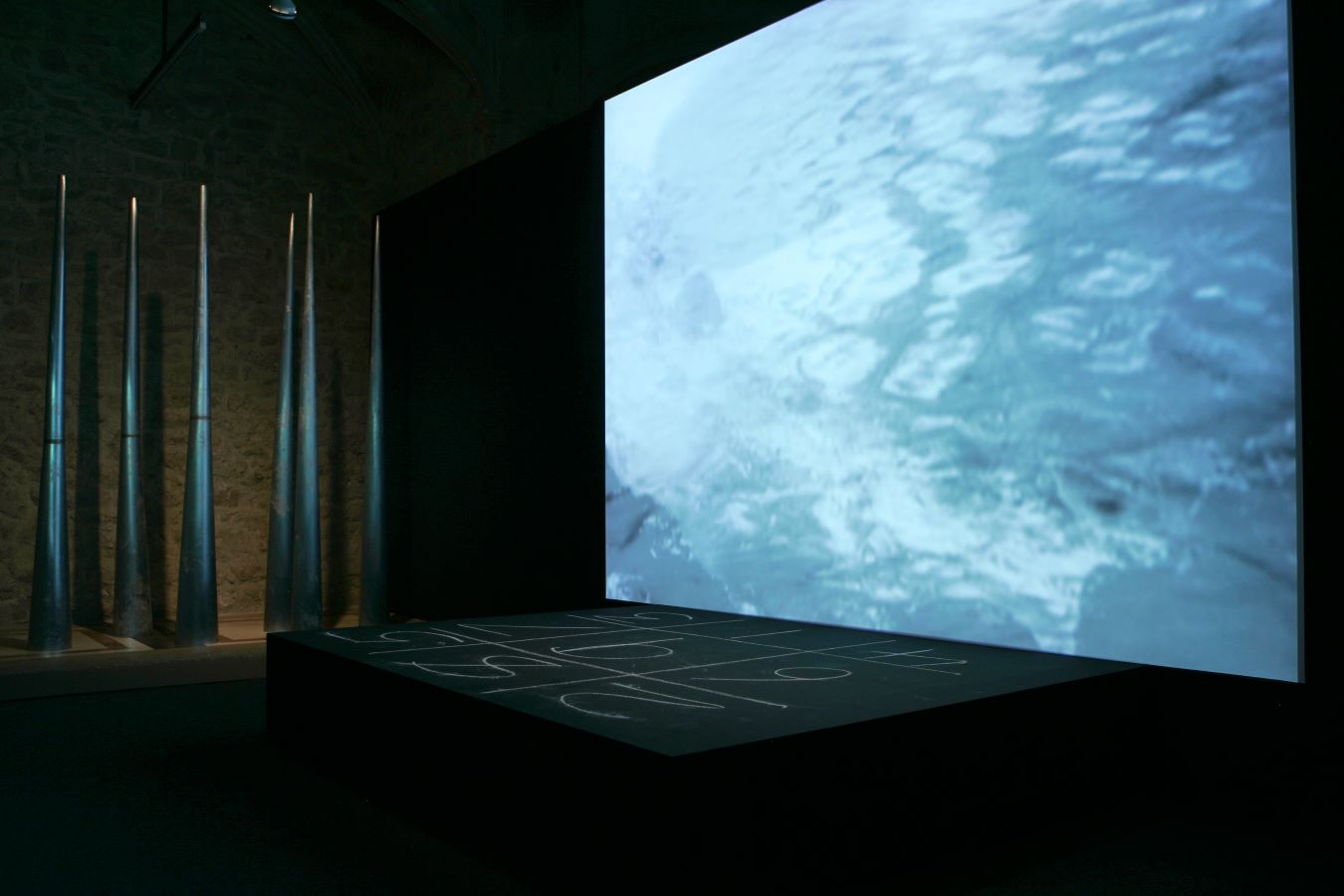 A video projection of water is above a small stage with a chalk number grid written on it. To the left are six large metal cones.