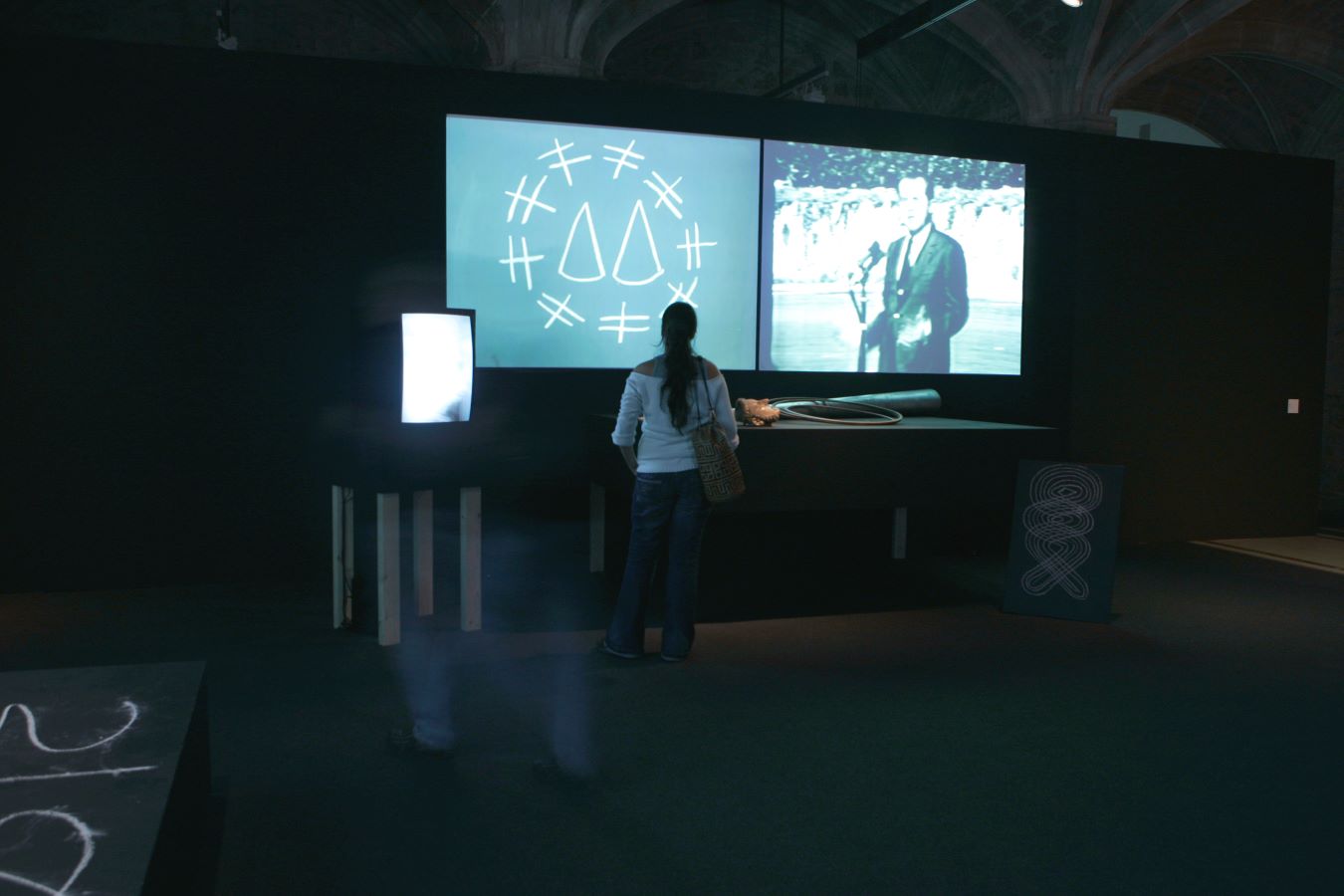 A figure stands in front of a table with various props on top of it. Behind the table are two video projections. To the left of the figure is a TV monitor on a stand; to the right is a chalk drawing. In the bottom left corner is a section of a small stage with a chalk number grid written on it.