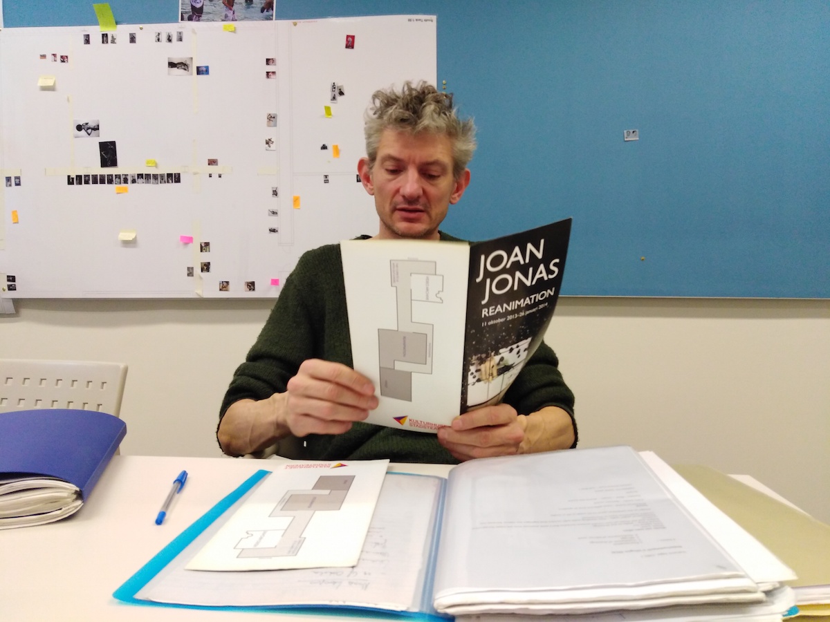 A man sitting behind a desk with a folder on top of it while reading a pamphlet titled: âJoan Jonas Reanimationâ.