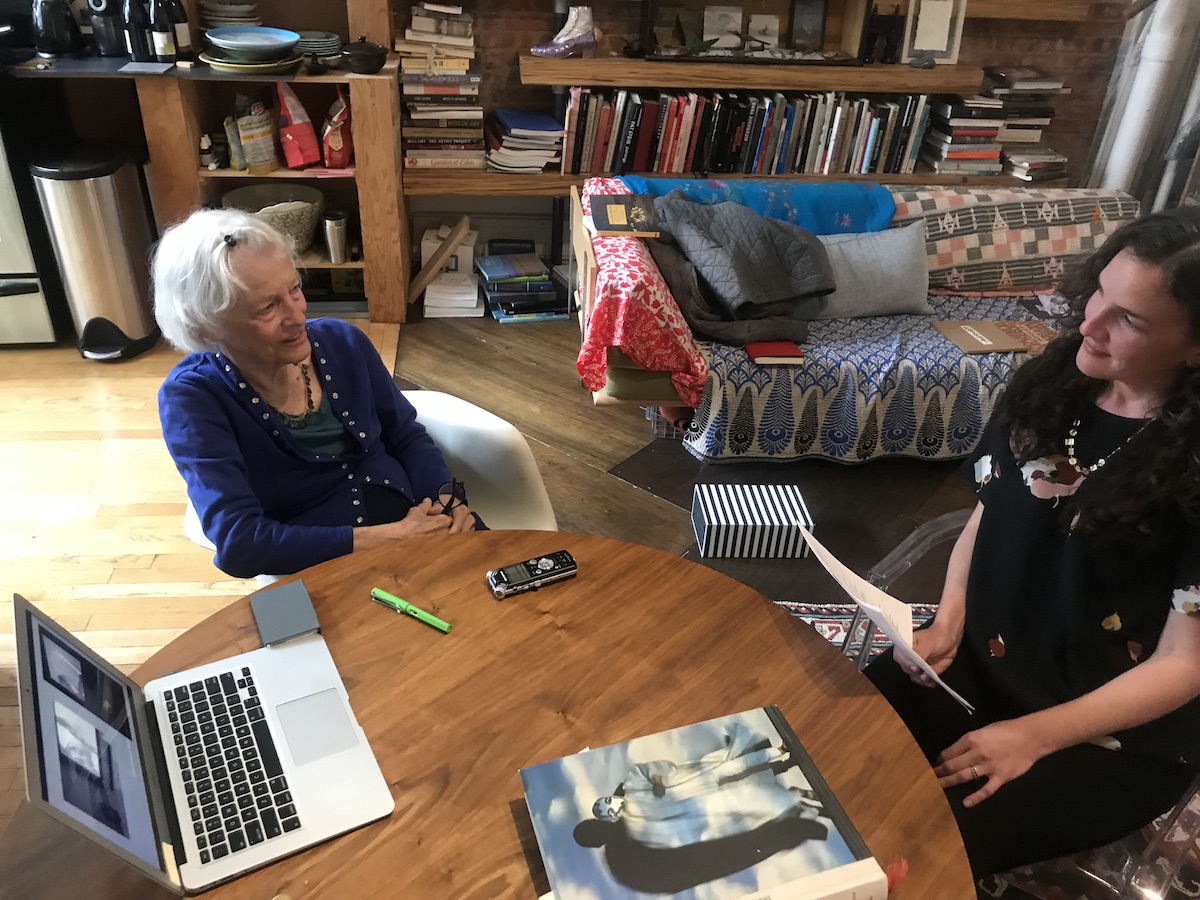 Two people sit at a table inside a home. On the table is a recording device, a laptop, and a book.