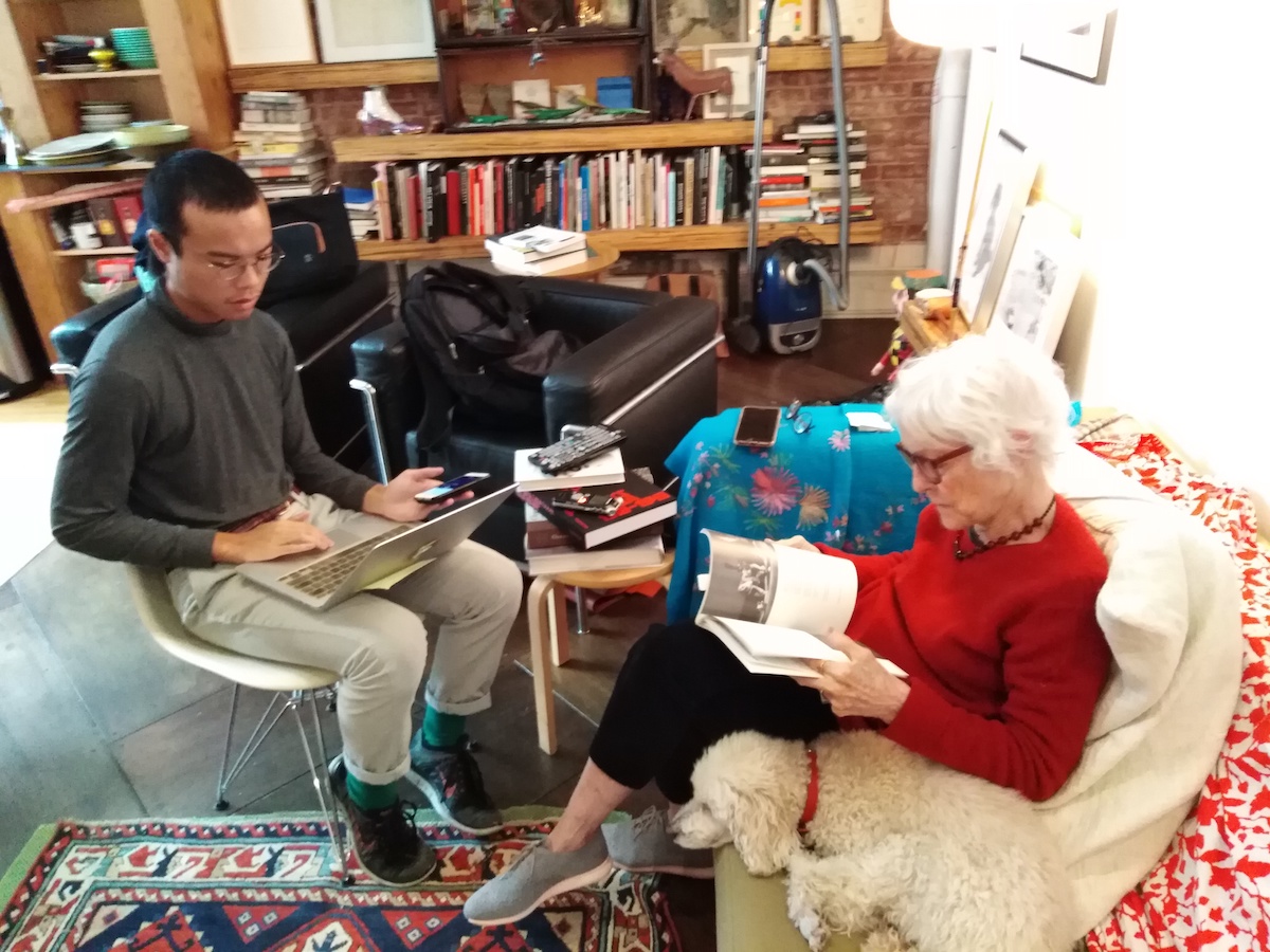 A man (left) sitting in a chair with a laptop next to a woman (right) who is sitting on a sofa and flipping through a book, next to the woman is a dog.