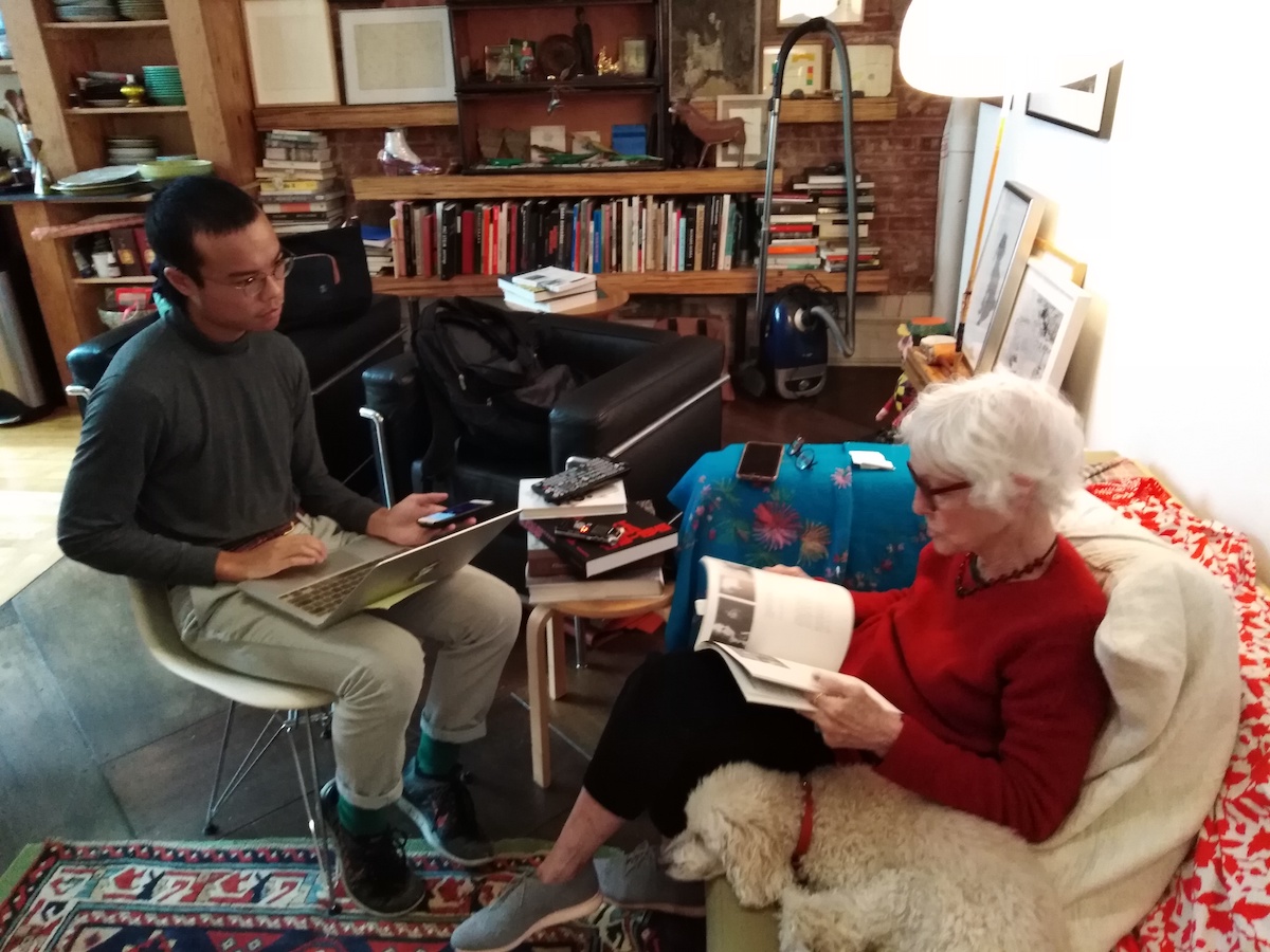 A man (left) sitting in a chair with a laptop next to a woman (right) who is sitting on a sofa and flipping through a book, next to the woman is a dog.