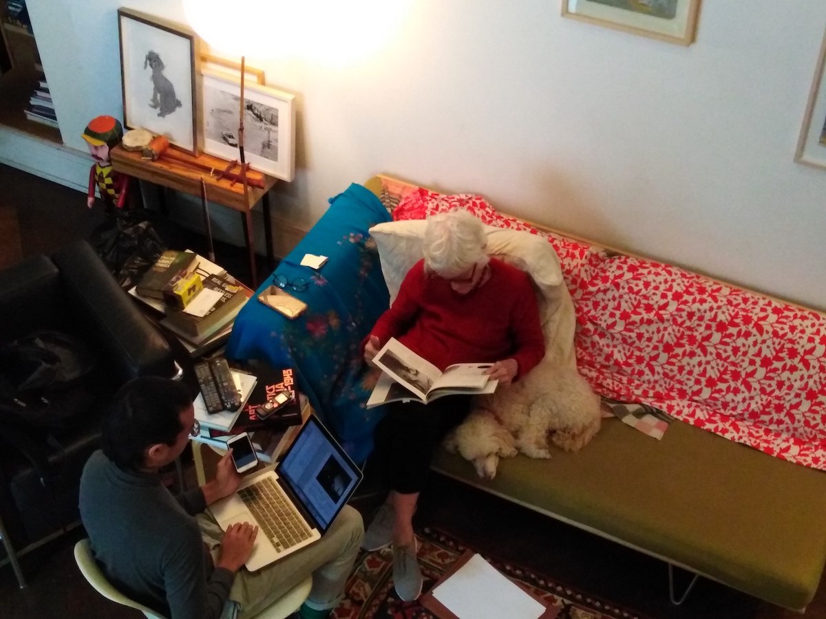 An aerial perspective of a man (left) sitting in a chair with a laptop next to a woman (right) who is sitting on a sofa and flipping through a book, next to the woman is a dog.