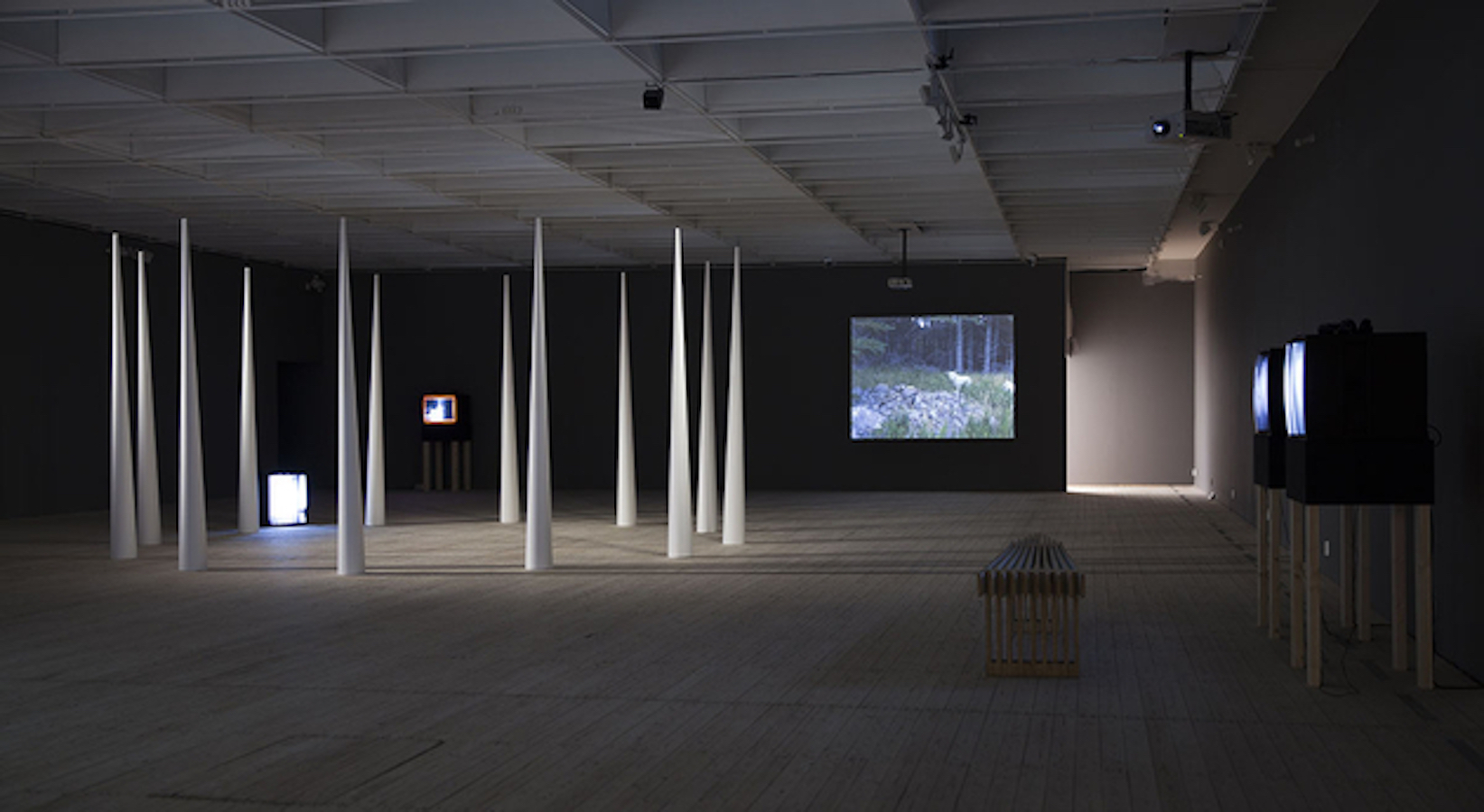 A room filled with three TV monitors on stands and one monitor on the floor, a video projection against the back wall and a circle of large cones.