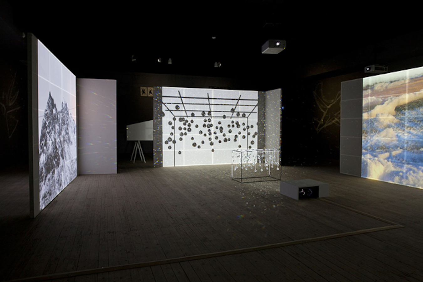 A sculpture made out of crystals is placed in front of a projector in the middle of the floor, surrounding it are three screens with video projections.