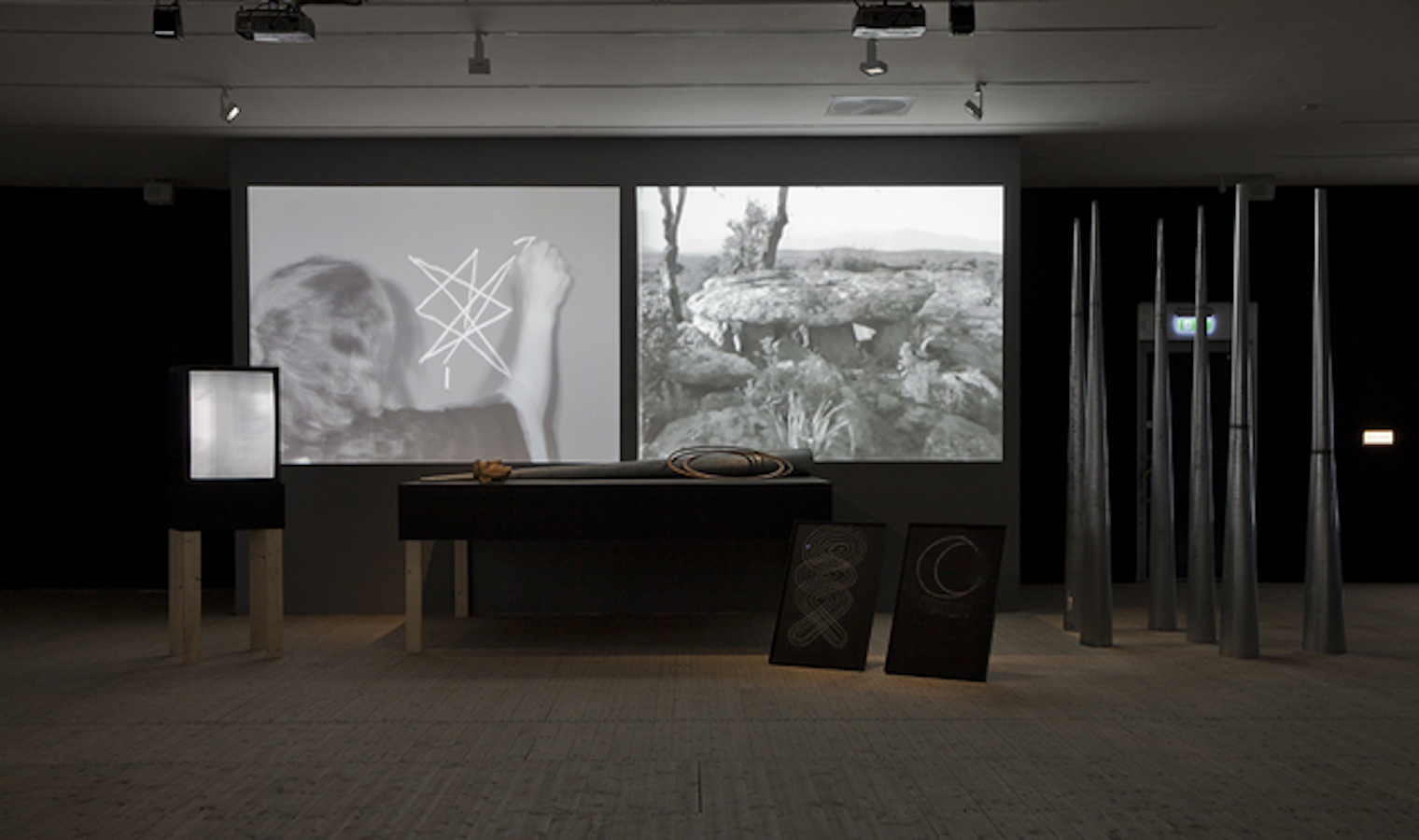 Various props used for Jonas’s performances, including: a TV monitor on a stand; a cone, hoop and mask on top of a table; two chalk drawings and a group of large cones. Behind the table are two video projections: one of Jonas drawing with chalk and the other showing a desert landscape.