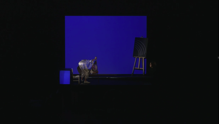 Jonas on stage bent over in front of a blue projection, holding a stick. There is a chalk drawing placed on an easel and a floating head to the right, and a TV monitor to the left.