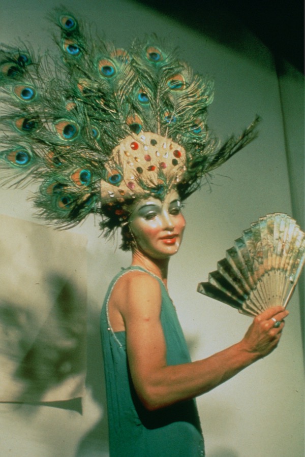 Jonas wearing a peacock-feathered headpiece, mask, and green dress while holding a fan