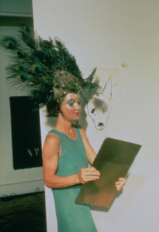 Jonas standing next to a drawing of a dog’s head while holding a mirror and wearing a peacock-feathered headpiece, mask, and green dress