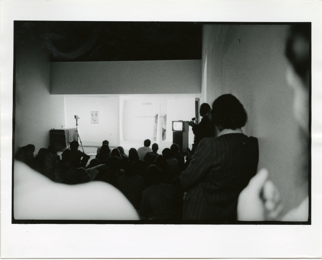 Audience faces towards a camera on a tripod, a drawing of a dog’s head, a video monitor on a stand, and a white cone.