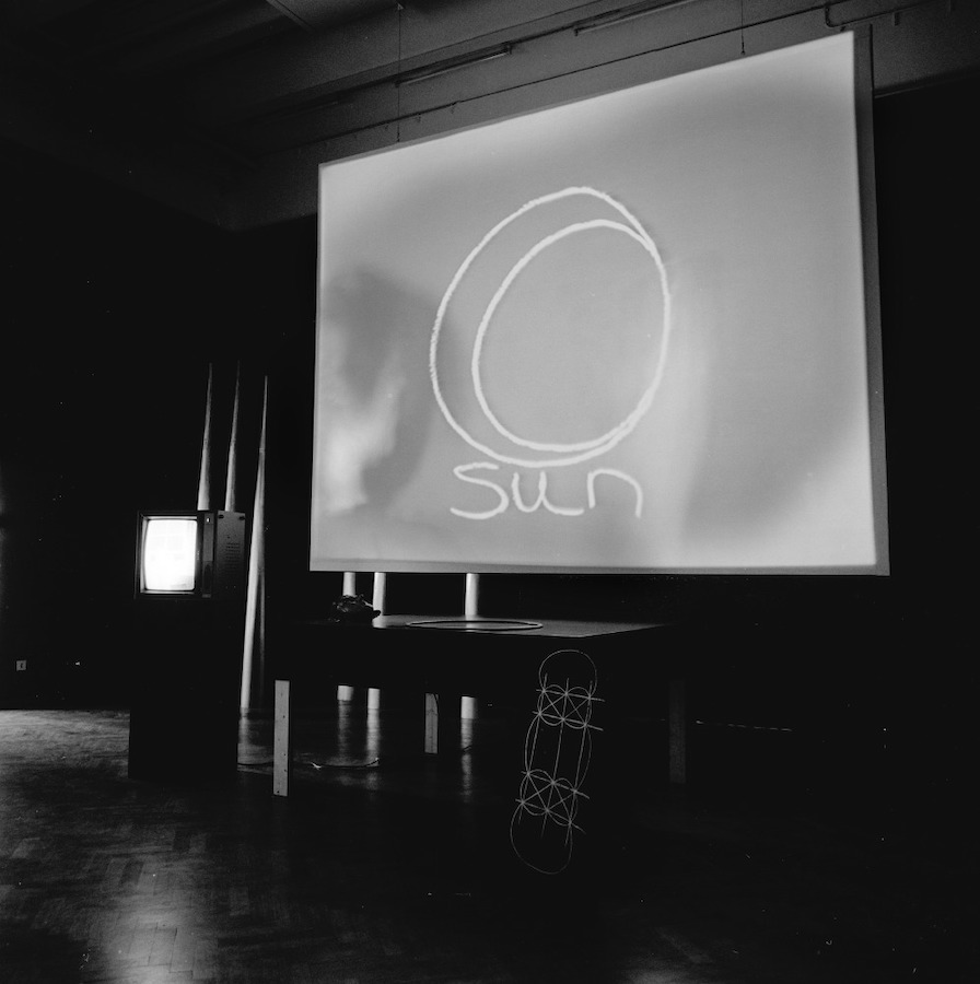 Various props used for Jonas’s performance, including: large cones, a TV monitor on a stand, a table with a hoop and mask, a drawing, and a projection of a circle drawing with the word “Sun” written underneath.