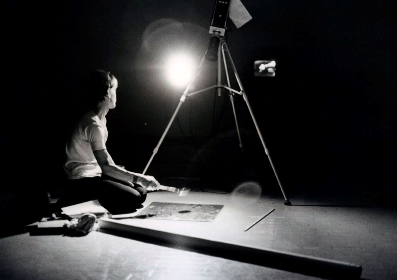 Jonas kneels below a camera on a tripod with a spoon in her right hand