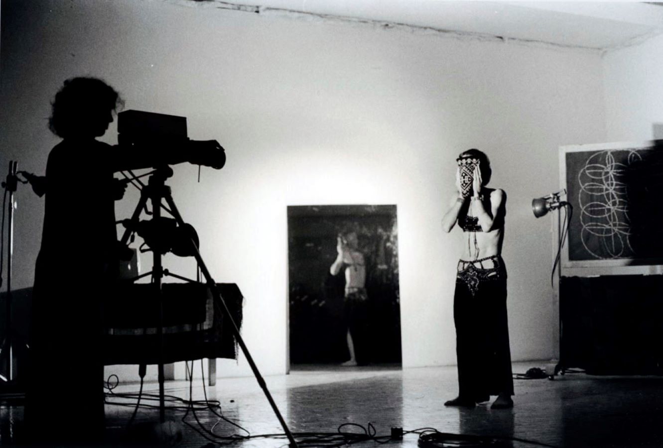 A camerawomen stands behind a tripod while Jonas covers her face. To the left, is a large mirror reflecting Jonas.